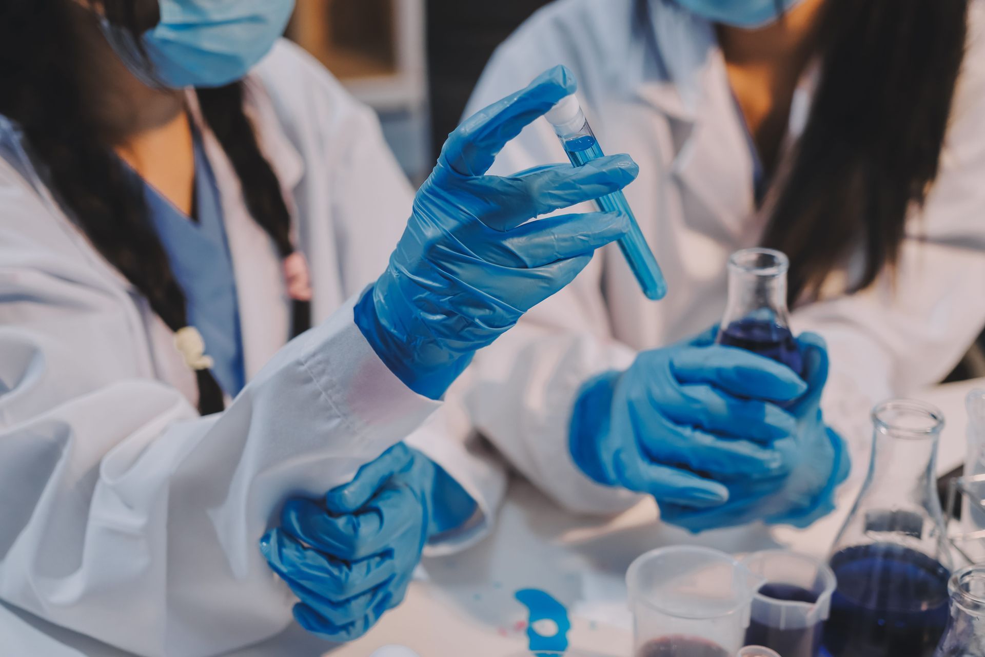 Two people in white lab coats and blue gloves examine a blue liquid in a test tube inside a laboratory.