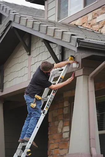 Person on a ladder using a power drill to repair the exterior of a house.