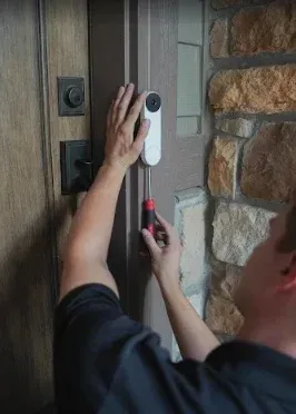 Person using a screwdriver to install a white doorbell/intercom on a wooden door next to a stone wall