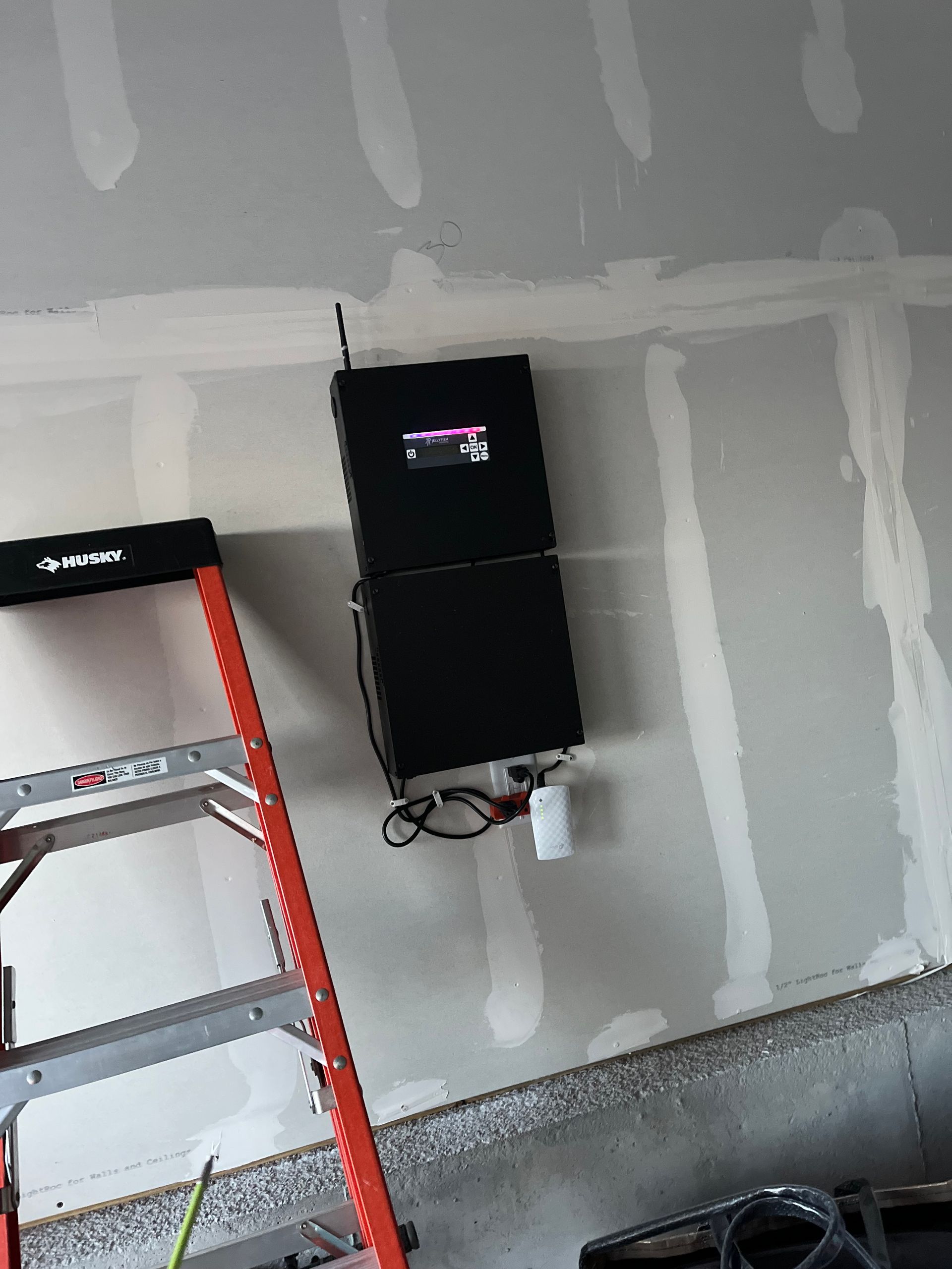 Garage wall with mounted black electrical unit, exposed wires, and a ladder on the left.