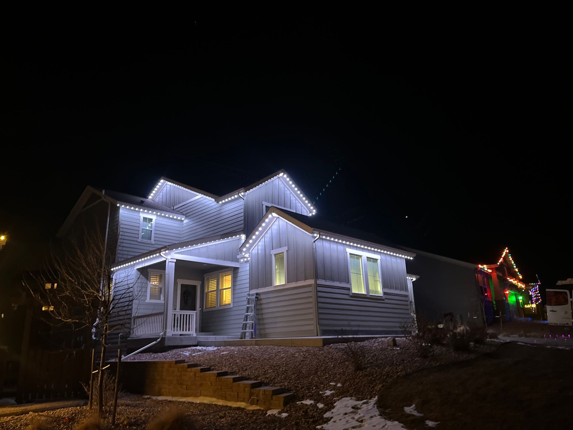 Snowy house at night decorated with white roof lights and warm lit windows.