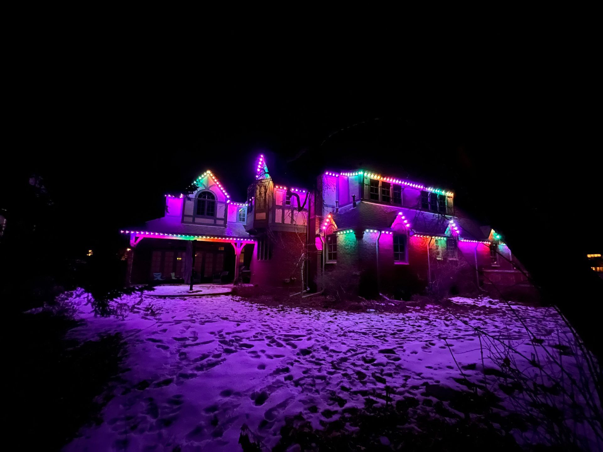 House outlined with colorful Christmas lights glowing purple, green, and pink at night on a snowy yard