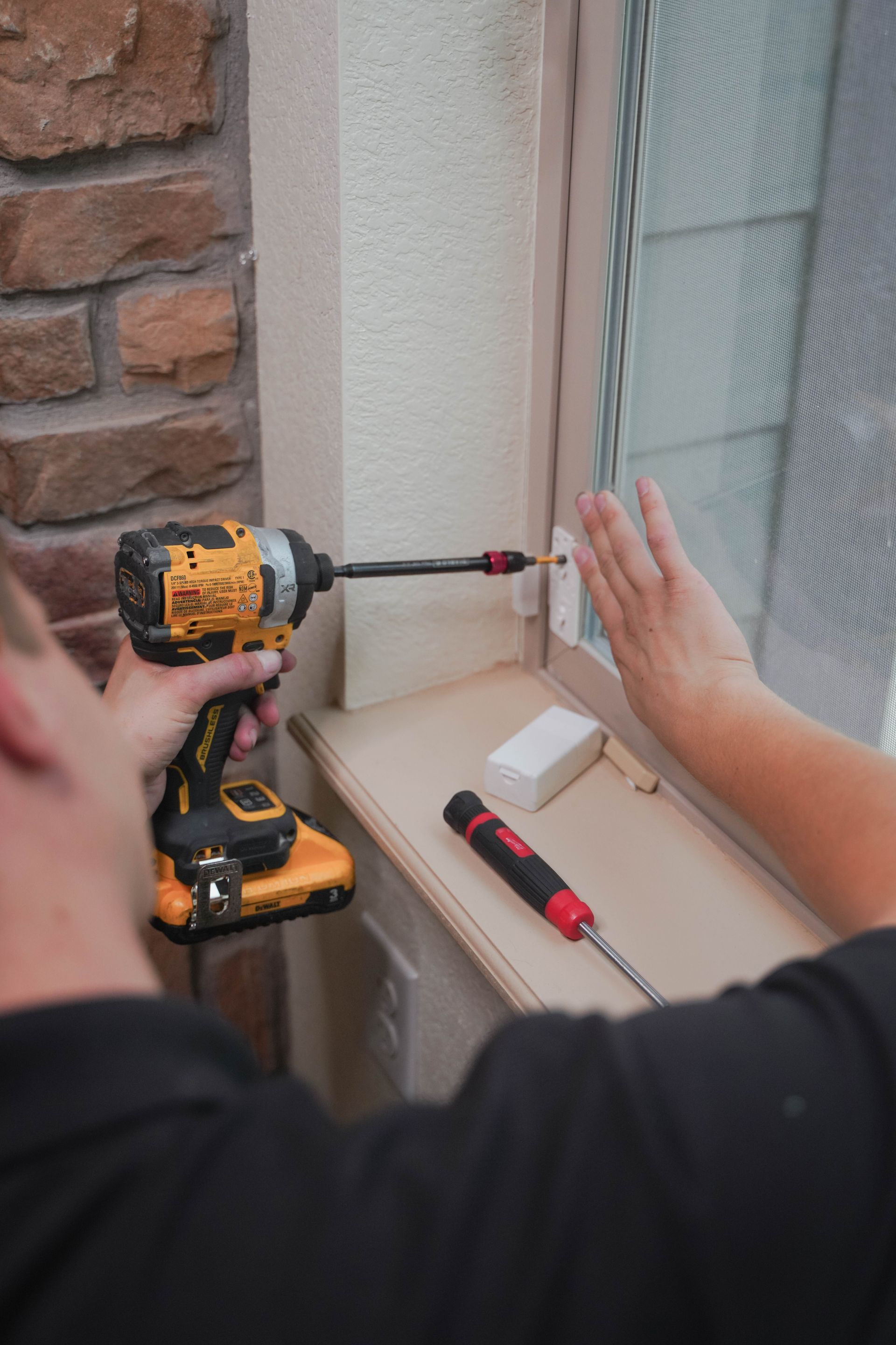 Person using a drill to install a screw beside a window frame on a brick wall.
