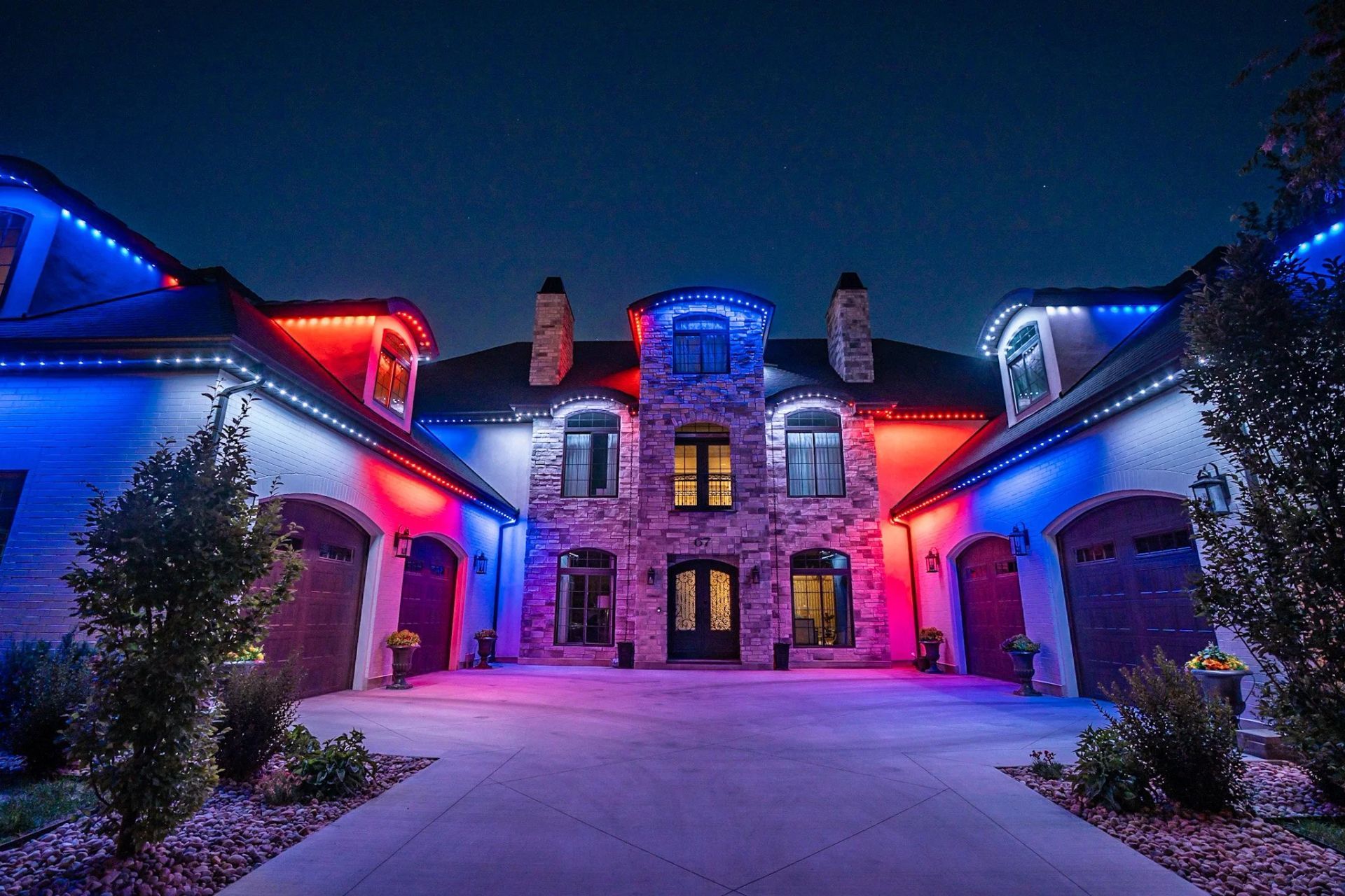 Large stone house at night, lit with blue and red holiday lights, with snow-covered driveway and trees