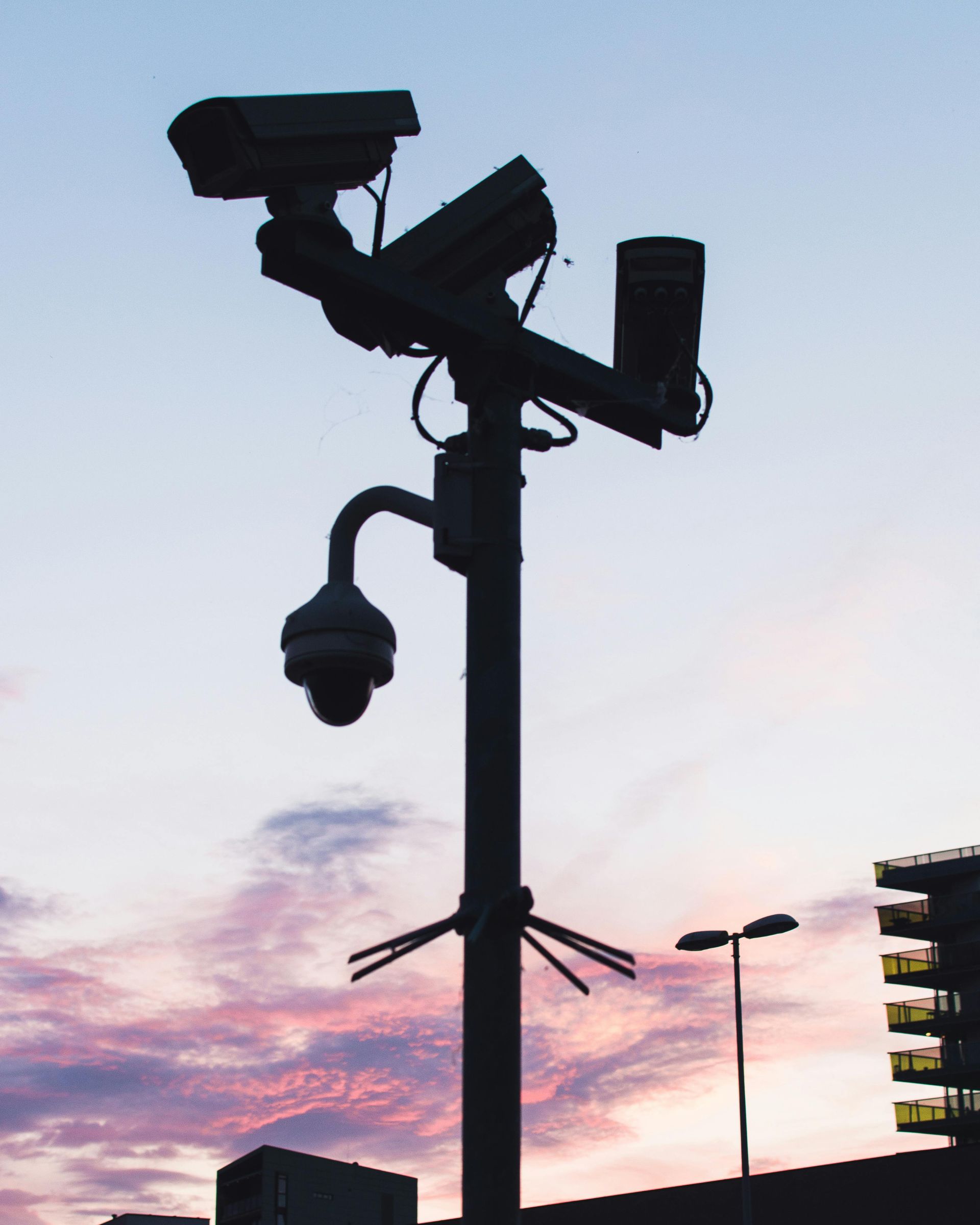 Silhouetted security camera pole at dusk against a pink-blue sky over buildings