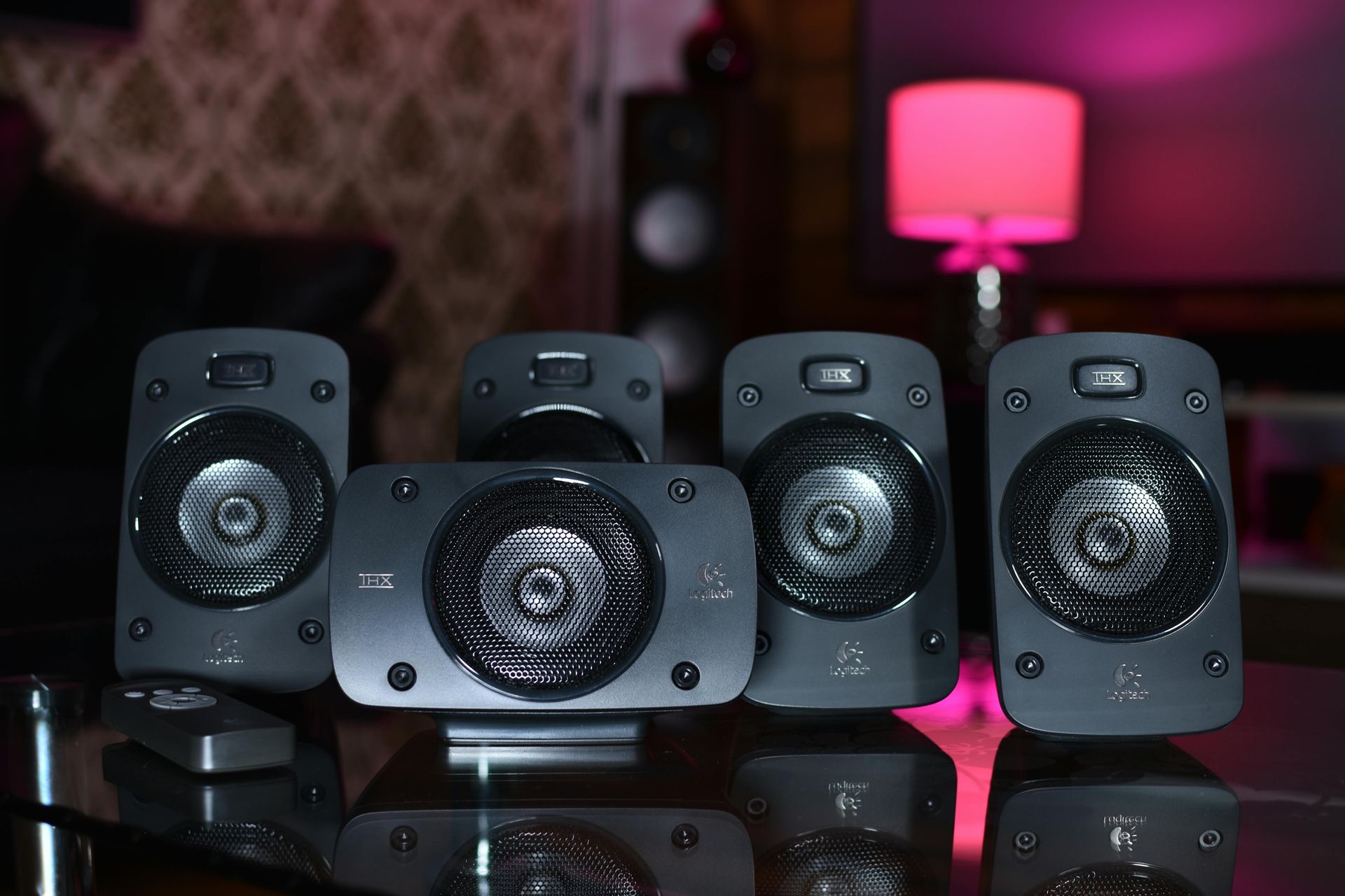 Five black speakers arranged on a reflective surface, with a pink lamp glowing in the background.