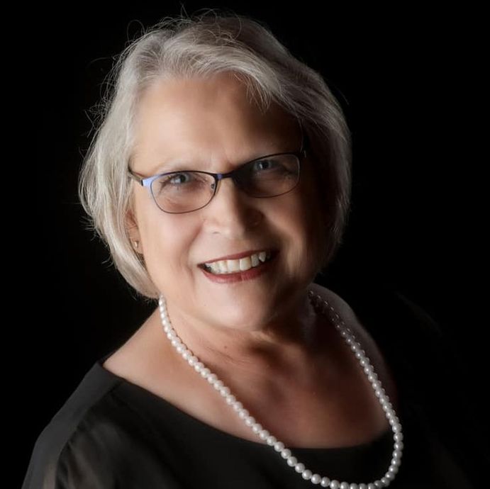 Smiling woman with gray hair, glasses, and pearl necklace against a black background