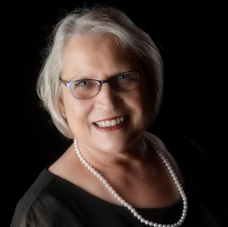 Portrait of a smiling woman with short gray hair, glasses, and a pearl necklace on a black background