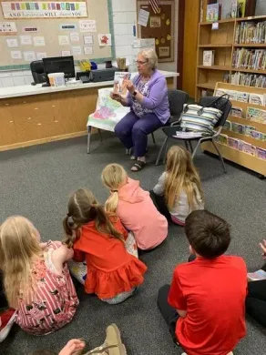 Children seated on a library carpet listening to a speaker in a bright room with bookshelves.