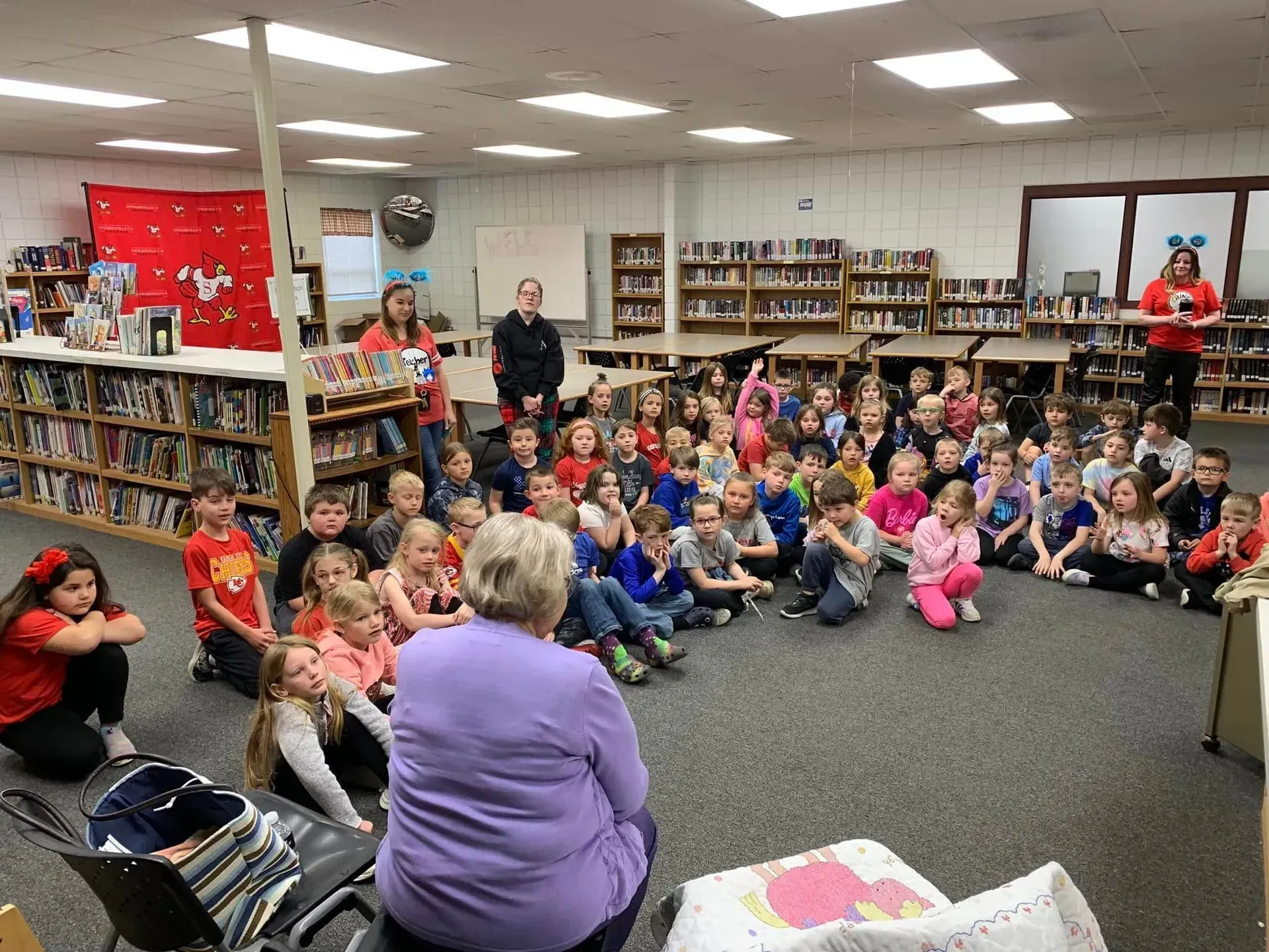 Children sitting on a library carpet listening to a speaker, with bookshelves and adults around them