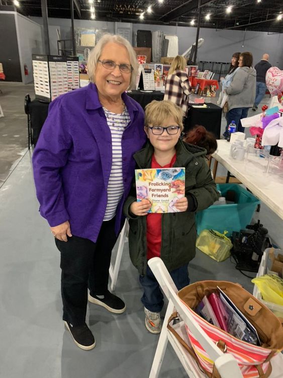 Two people smiling at a convention booth, one holding a colorful book or card.