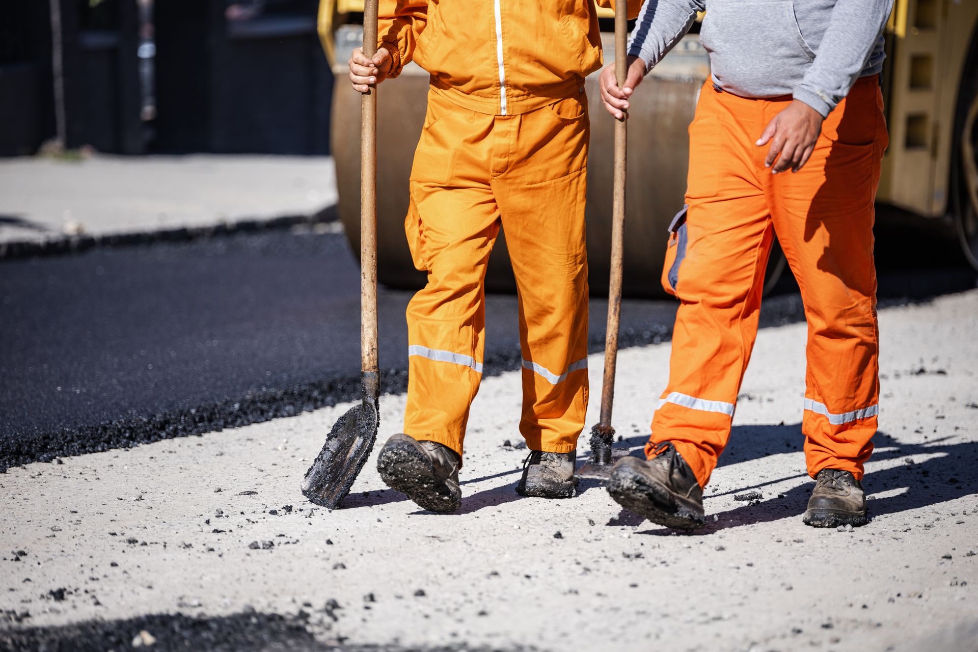Two workers in bright orange overalls hold tools while standing on a prepared section of a road. Two workers in bright orange overalls hold tools while standing on a prepared section of a road.
