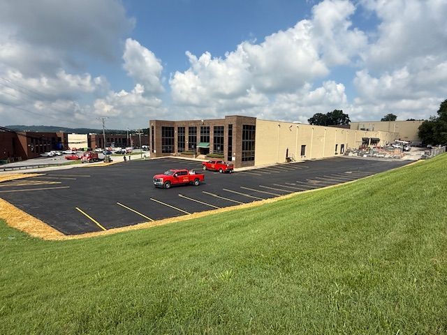 New commercial building with large parking lot, two red trucks, and green grassy hill under a cloudy sky.