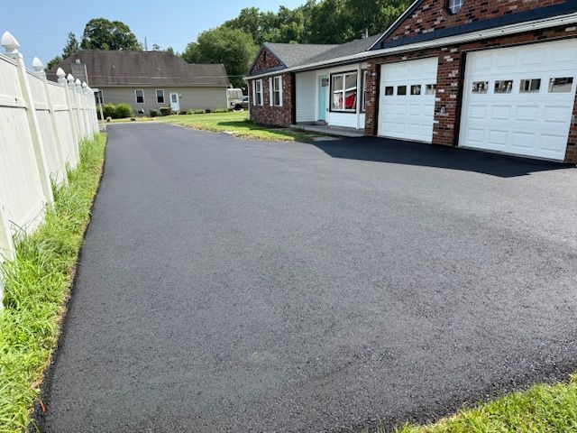Freshly paved asphalt driveway beside a house with white garage doors and a white fence