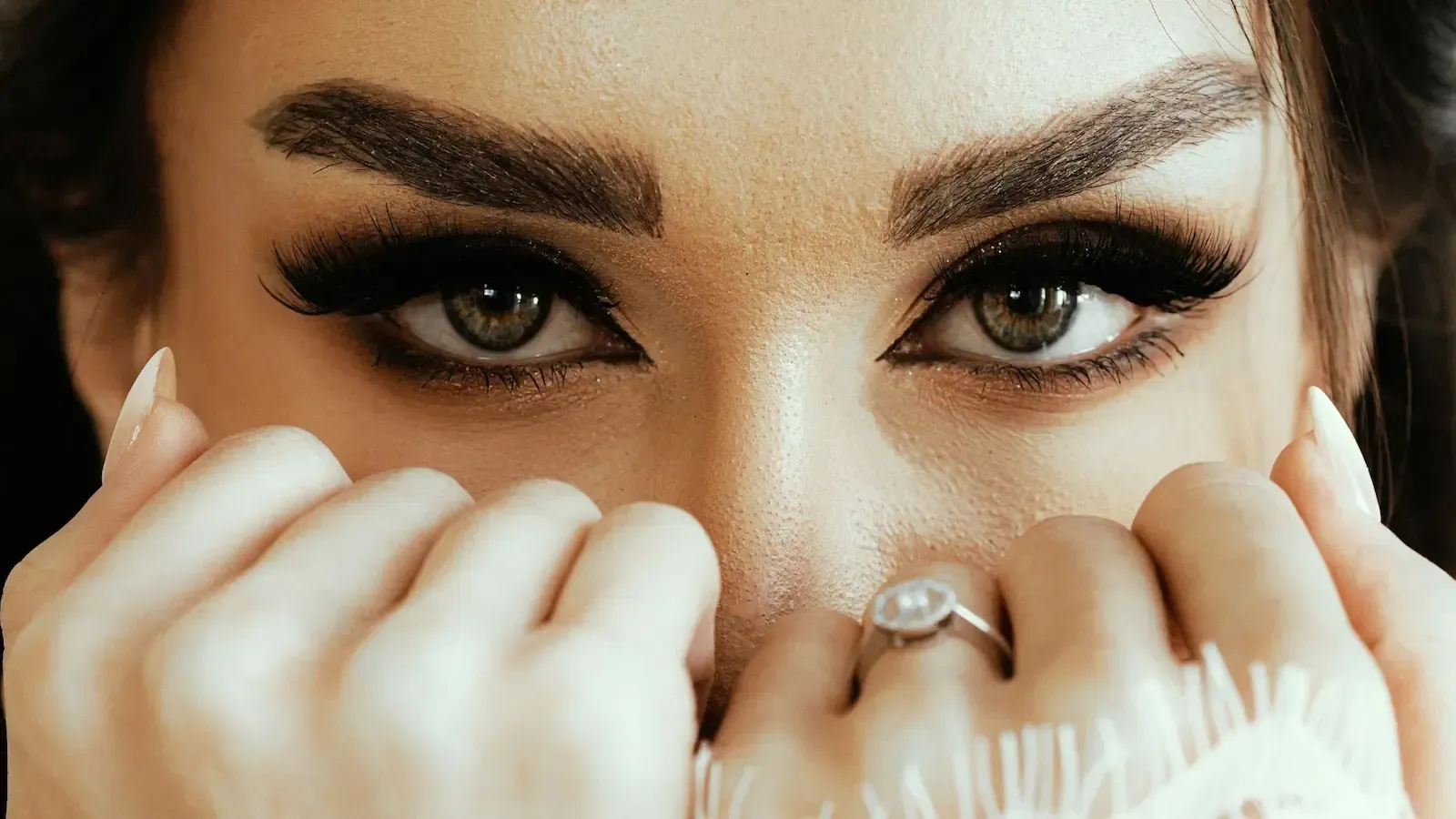 Close-up of eyes framed by dramatic eye makeup; hands partially cover face.
