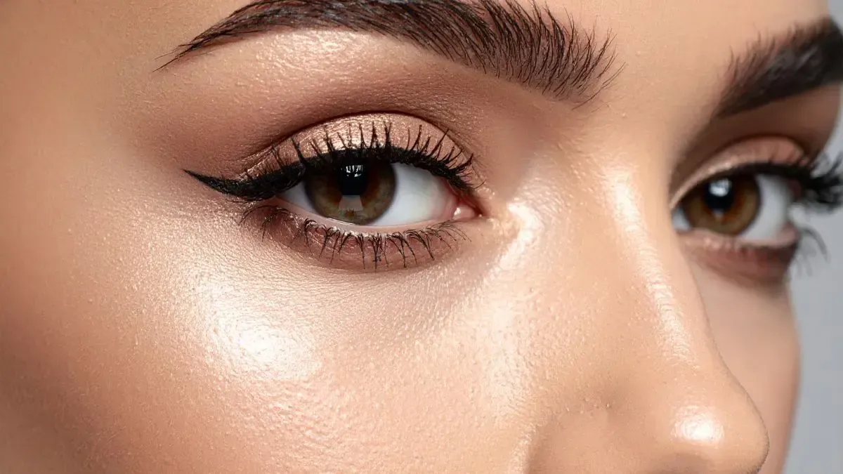 Close-up of a woman's face with brown eyes, winged eyeliner, and defined eyebrows. Her skin appears dewy.