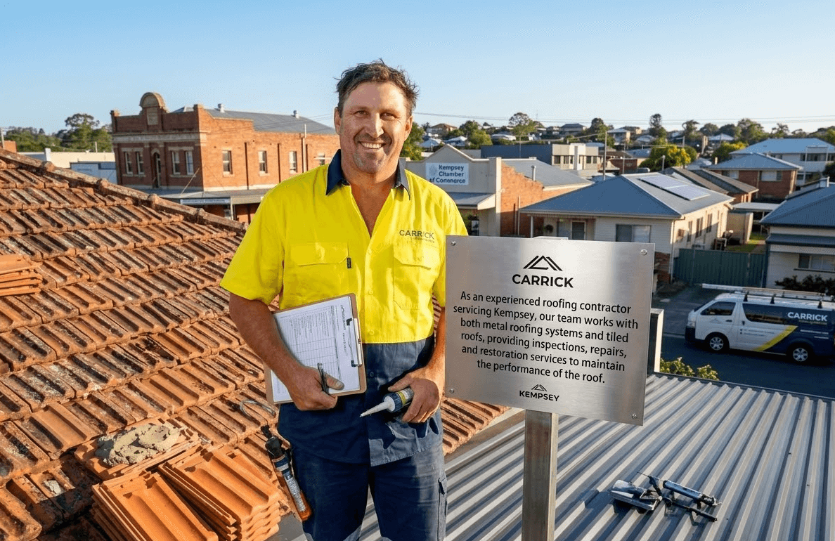 Man in yellow shirt on a terracotta tile roof, holding clipboard, smiling. Sign on roof. Buildings in background.