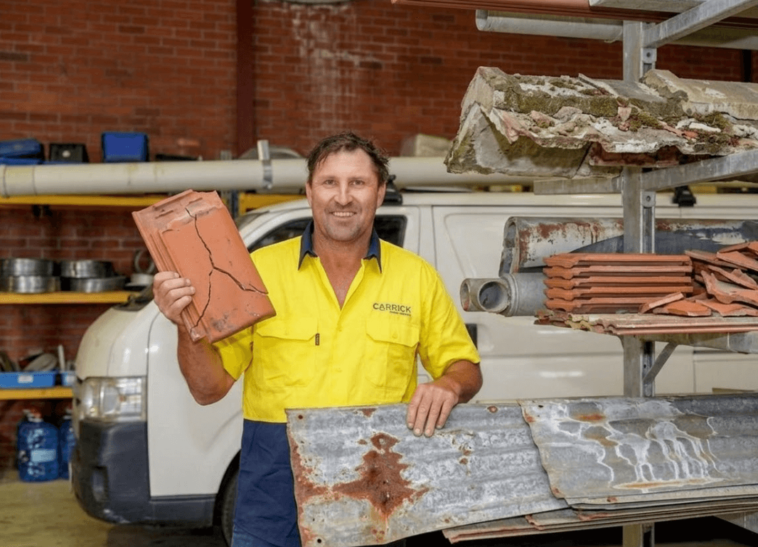 Man in yellow shirt holds broken roof tile, standing in front of van and shelves with roofing materials.