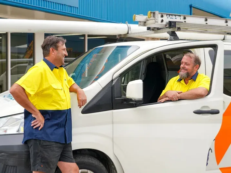 Two plumbers in yellow shirts talking beside a white utility van with a roof rack.