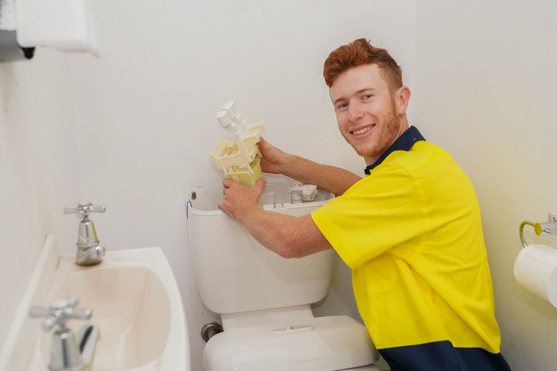 A man (Carrick Plumbing staff member) in a yellow shirt is fixing a toilet in a bathroom.