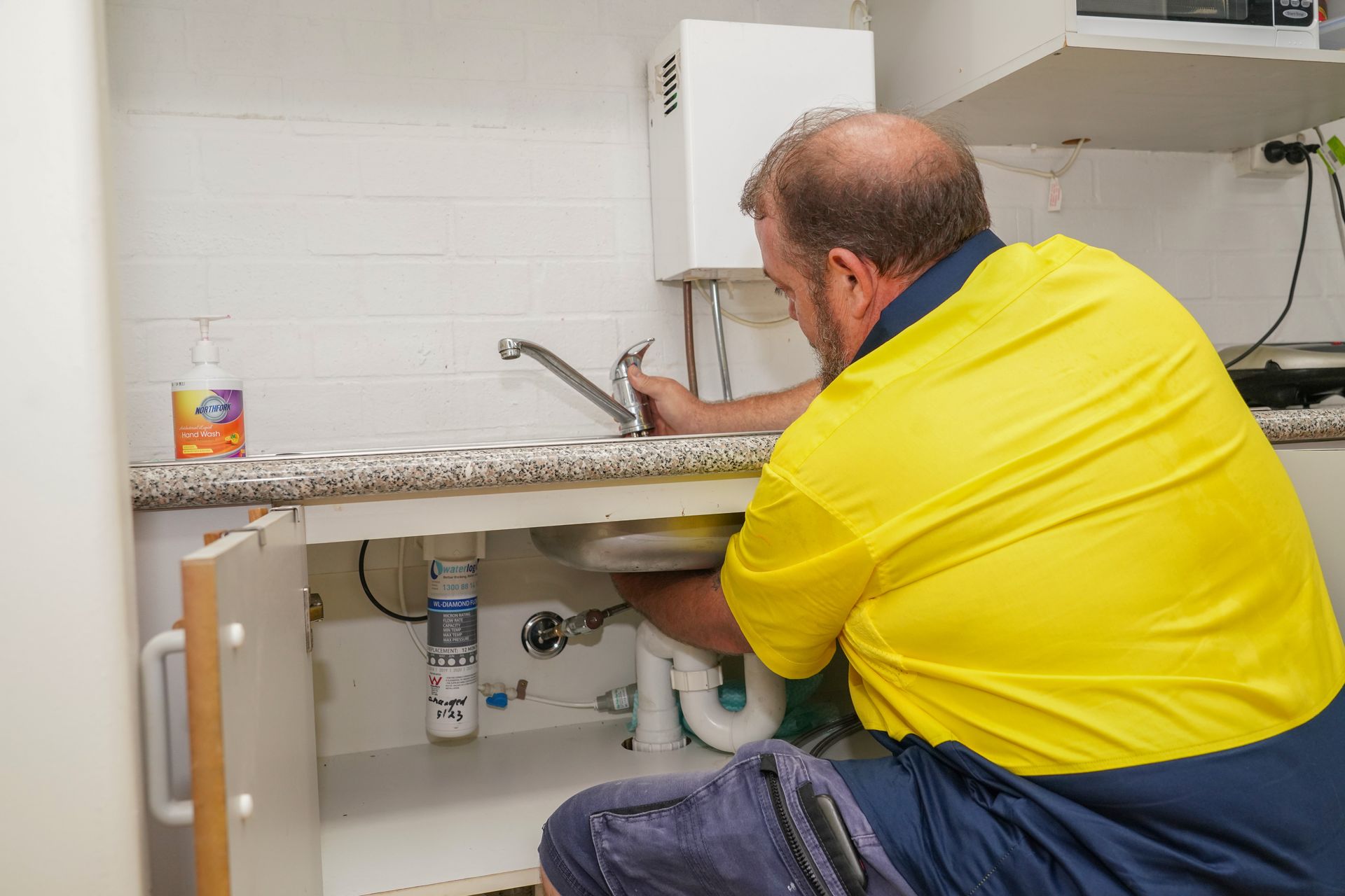 A person in a yellow high-visibility shirt performs plumbing work under a kitchen sink.