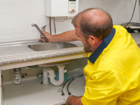 Carrick Plumbing team member in a yellow shirt is fixing a sink in a kitchen to a residential plumbing client.