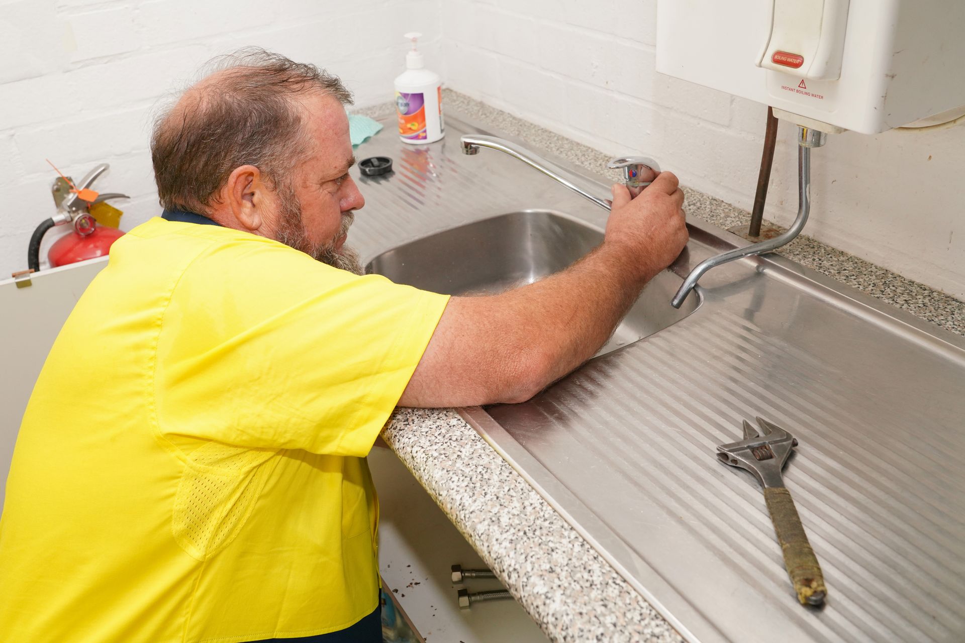 A person in a yellow uniform repairs a sink faucet, with an adjustable wrench lying on the stainless steel counter nearby.