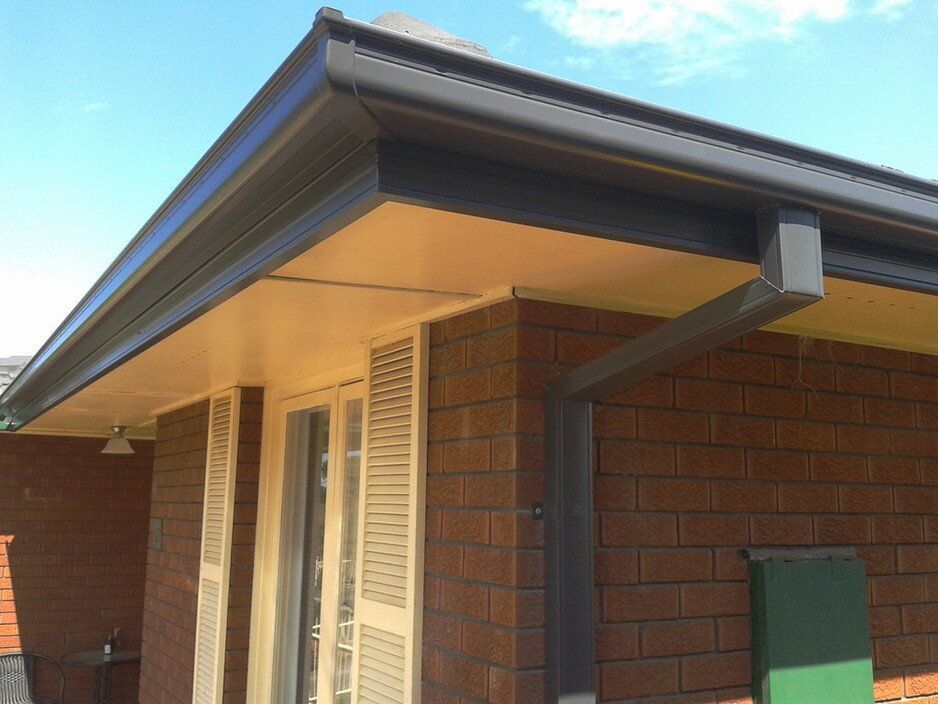 Dark gray gutters and downspouts attached to a brick house with a tan soffit under the roof eaves.