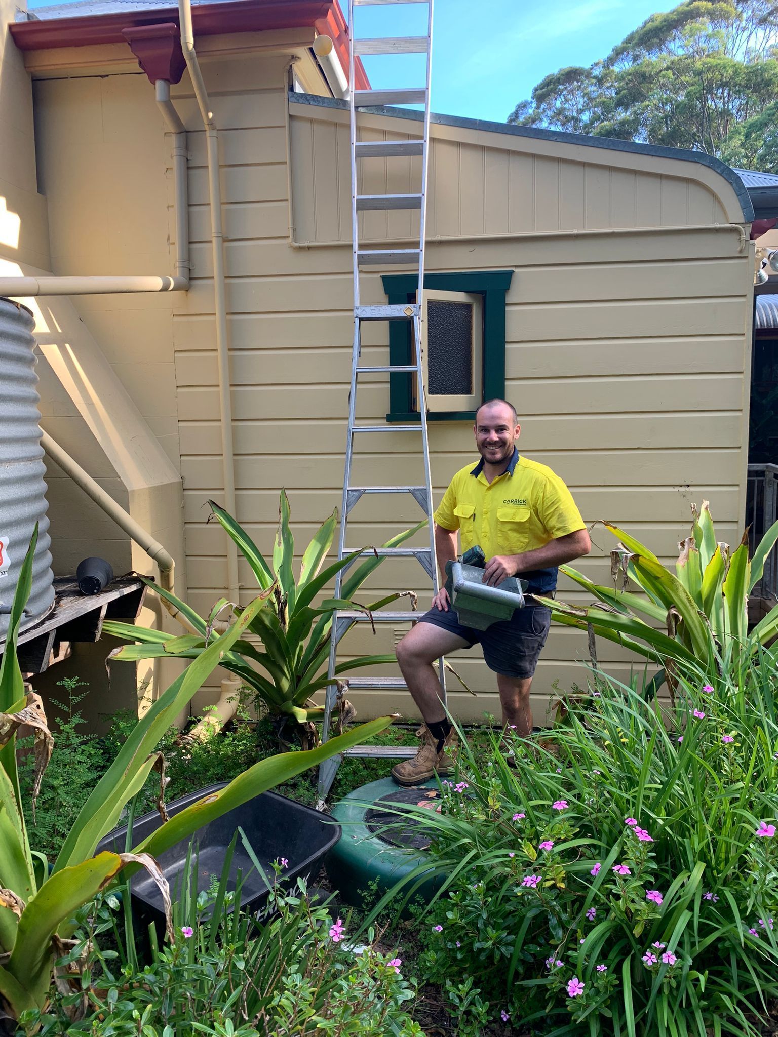 Matt a roofer from Carrick Plumbing standing on a ladder beside a yellow house with a garden below.