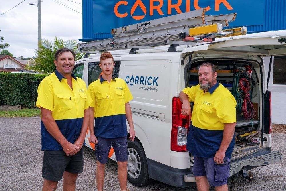 Three workers in yellow and navy uniforms stand in front of a Carrick branded service van with a roof ladder rack.