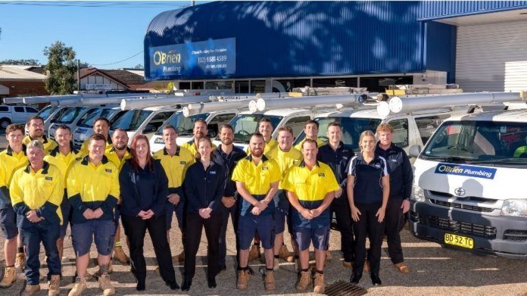 A group of 17 Carrick employees in yellow shirts and dark workwear standing in front of trade vans.