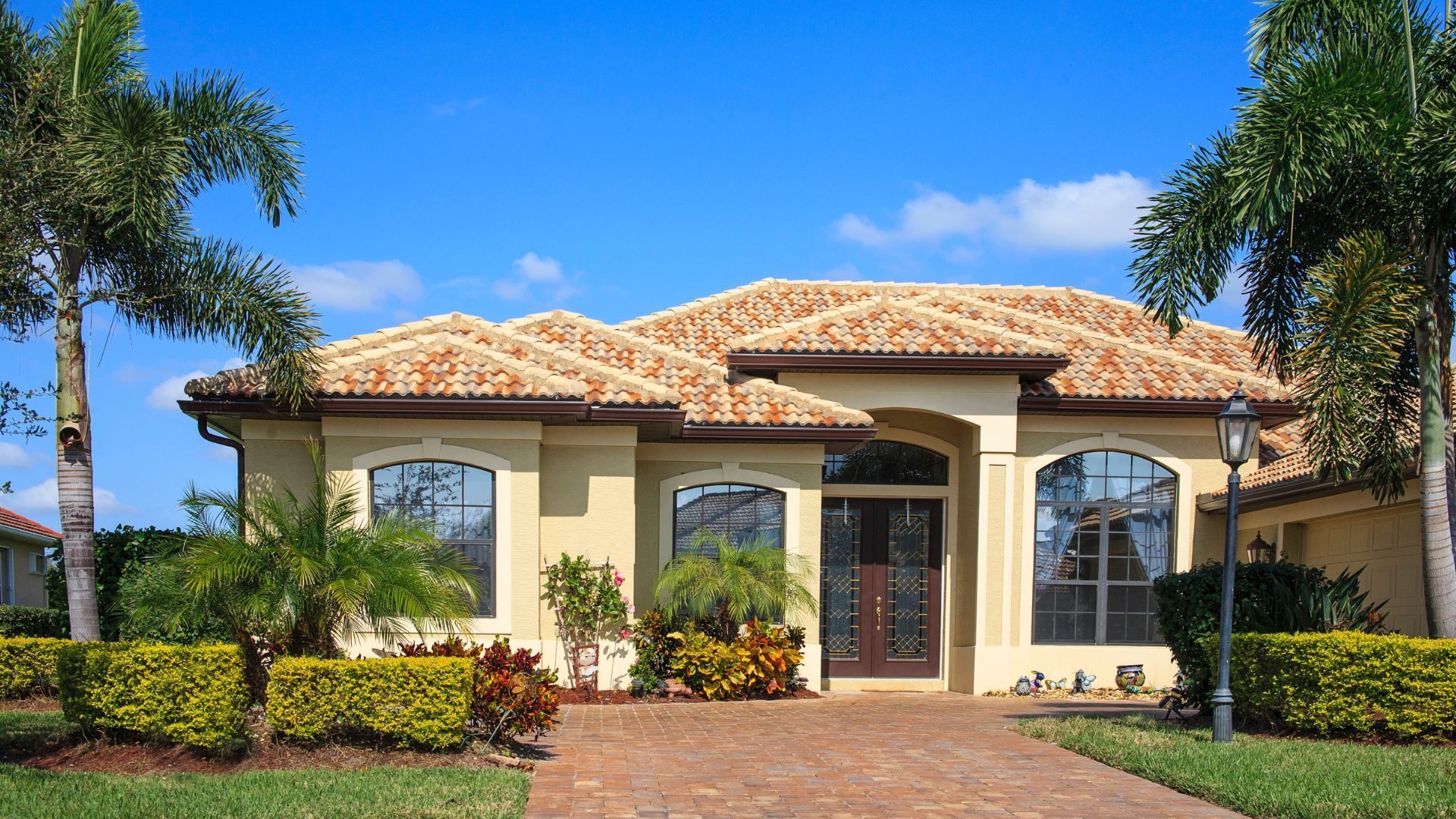 Mediterranean-style house with a tiled roof, arched entry, palm trees, and a brick driveway under a blue sky