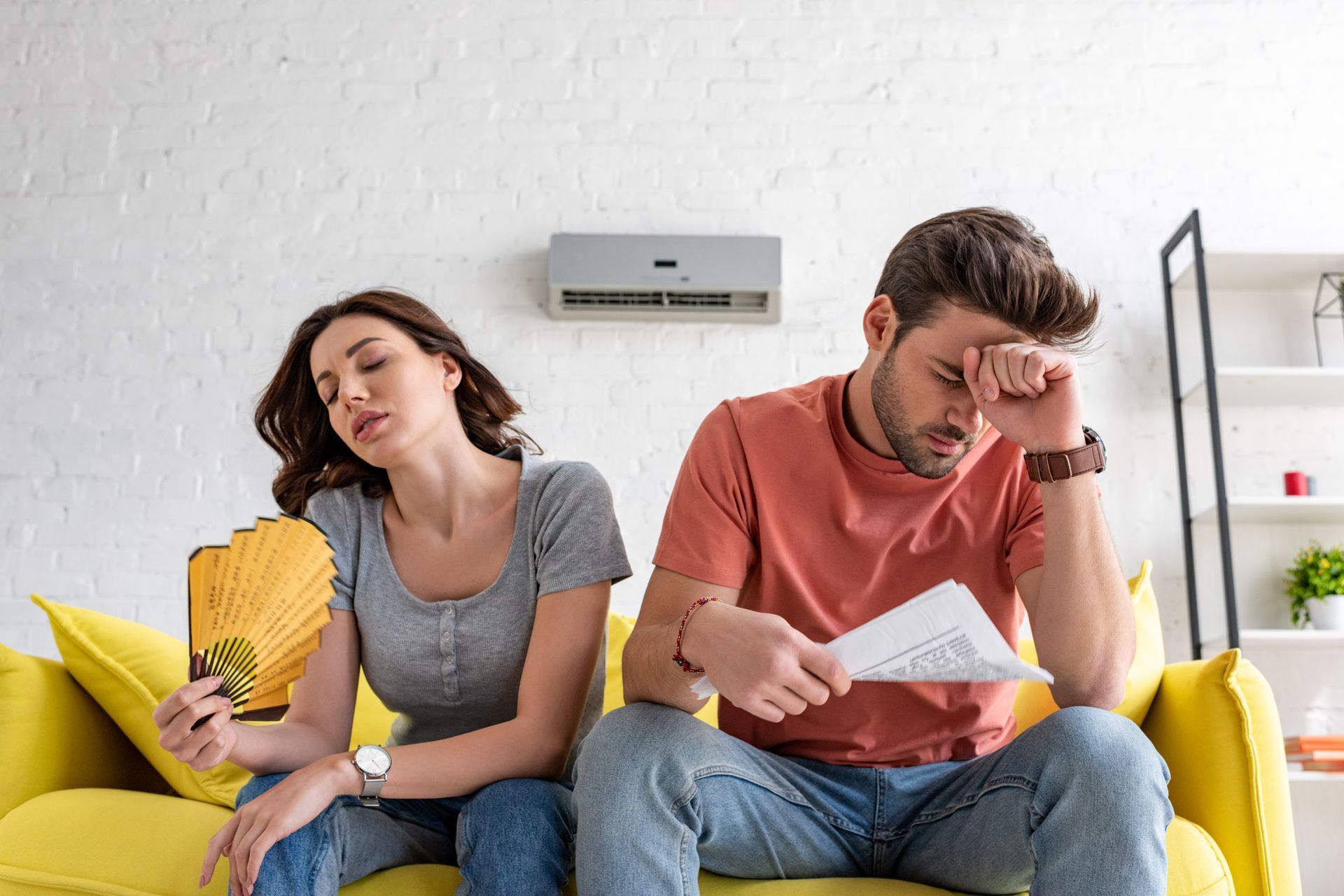 Two people sitting on a yellow couch, one reading a book and the other holding papers, looking stressed