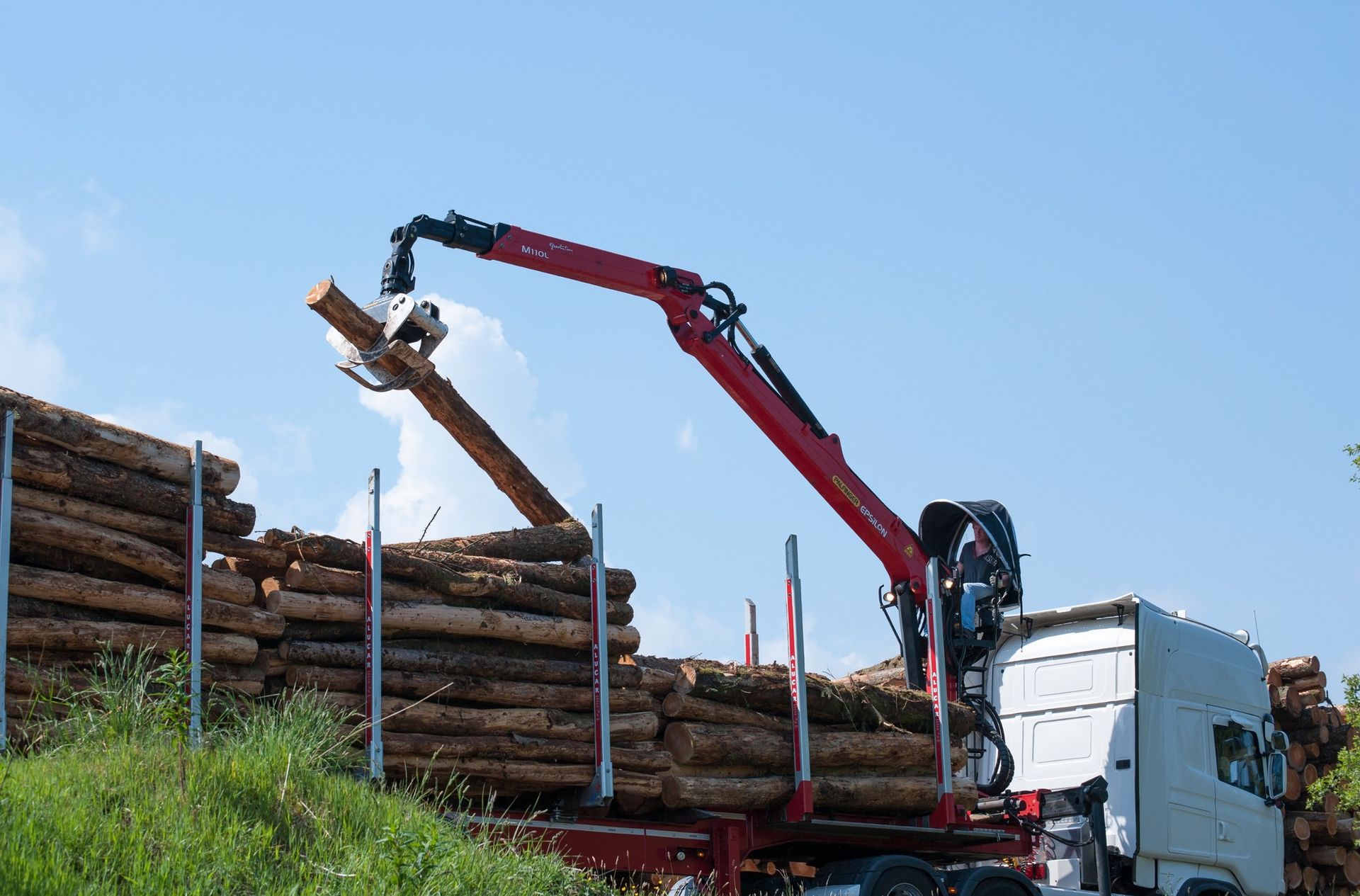 A logging truck uses a red crane to load logs onto its bed against a blue sky.