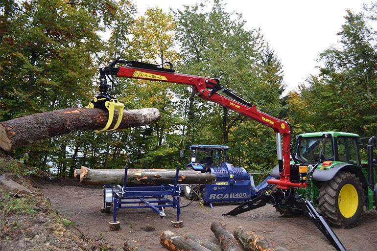 A tractor with a crane arm lifts a log; another log is on a saw. Trees in the background.