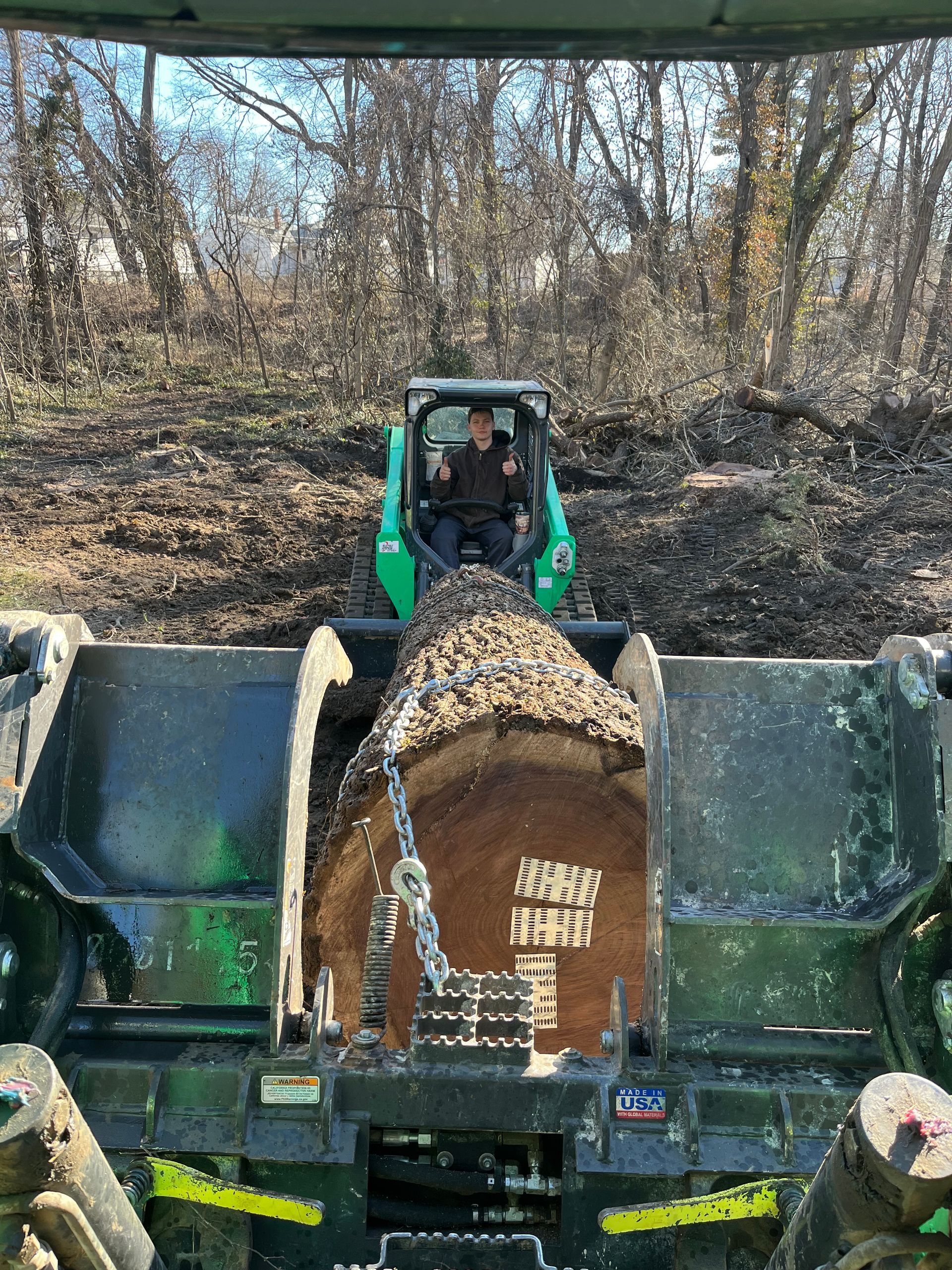 A person in a green Bobcat loader is holding a large log; sunny outdoor setting.