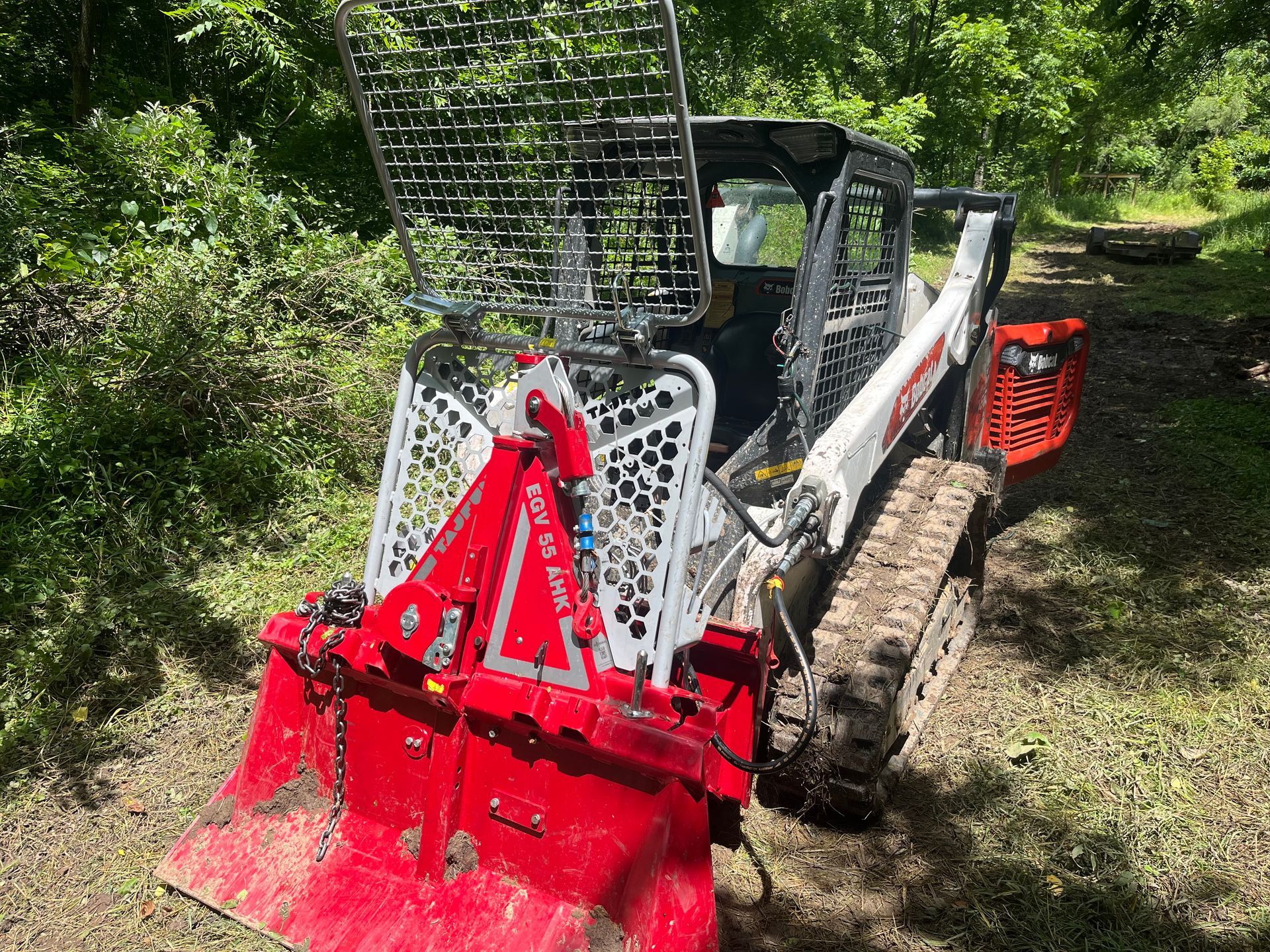 A white and black Bobcat skid-steer with a red mulcher attachment clearing brush in a wooded area.
