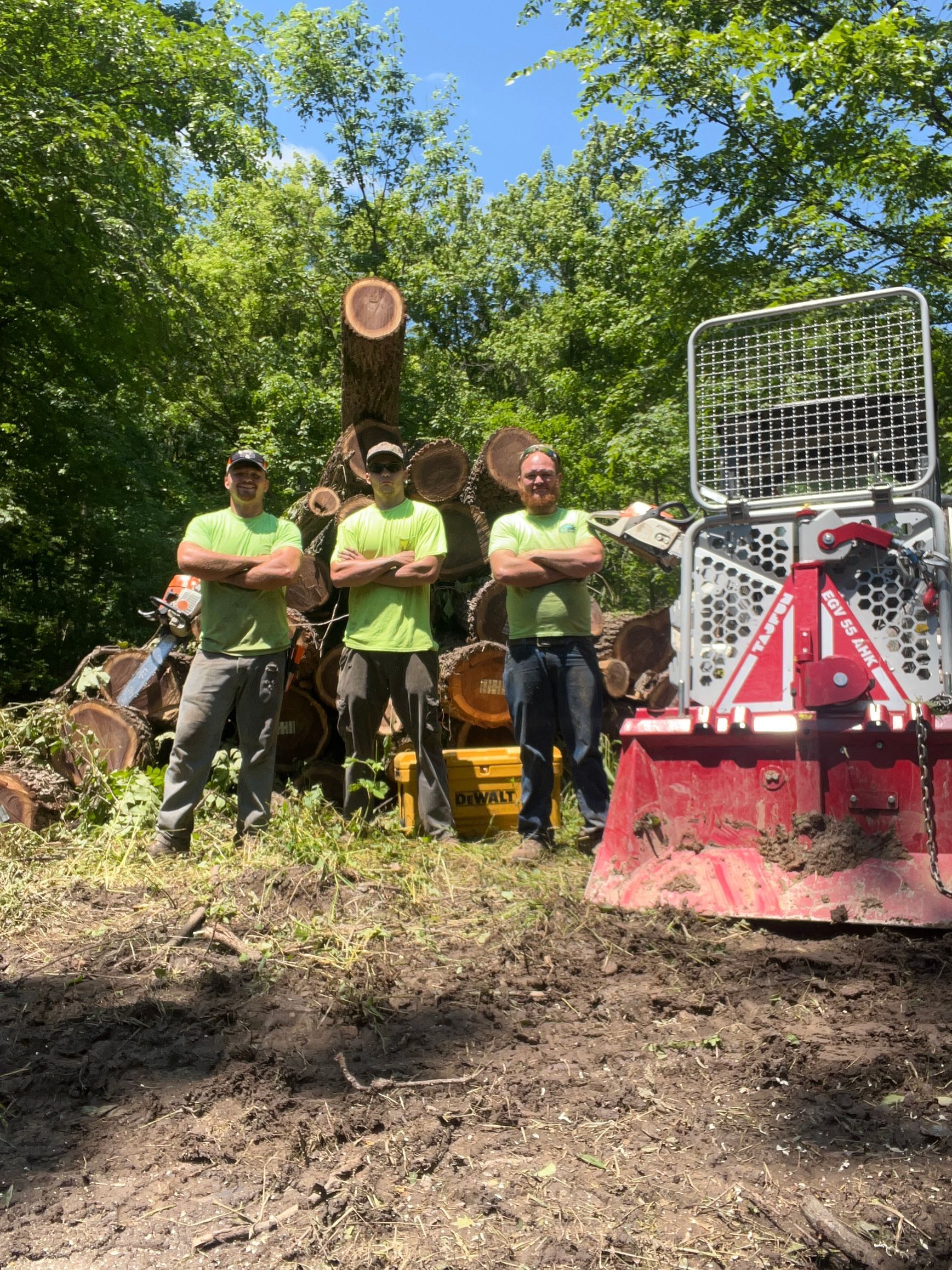 Three men in green shirts stand with arms crossed near logs and logging equipment in a wooded area.