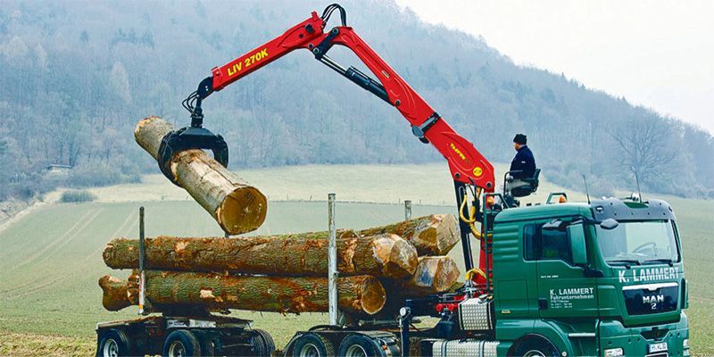 A logging truck loading logs. A red crane lifts a log into the truck bed. The truck is green, a man operates the crane.