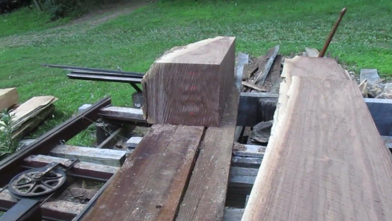 Wooden beam being sawed on a sawmill, with cut planks and grass background.