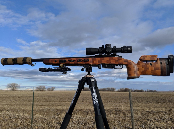 Rifle on a tripod in a field. The rifle has a camouflage pattern and a scope. Blue sky and clouds in the background.
