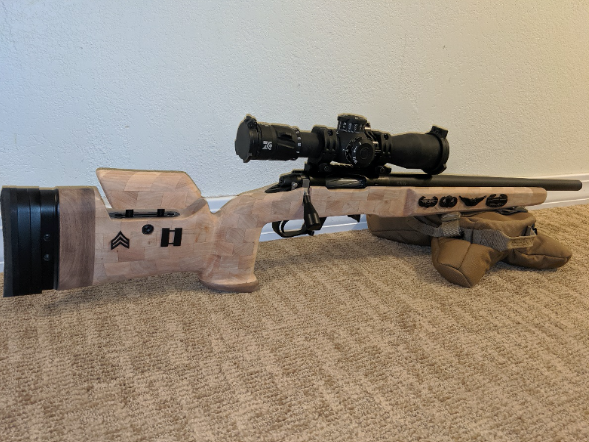 A bolt-action rifle with scope and wood stock, resting on a sandbag on a carpeted floor.