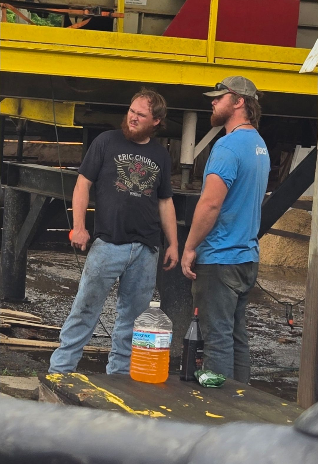 Two men in work clothes standing near a structure, one with a beard, a bottle of liquid is in front of them.