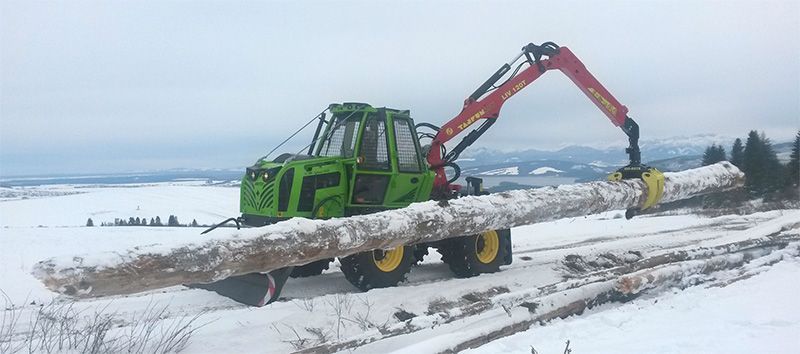 Green logging machine carrying a large, snow-covered log in a snowy, outdoor setting.
