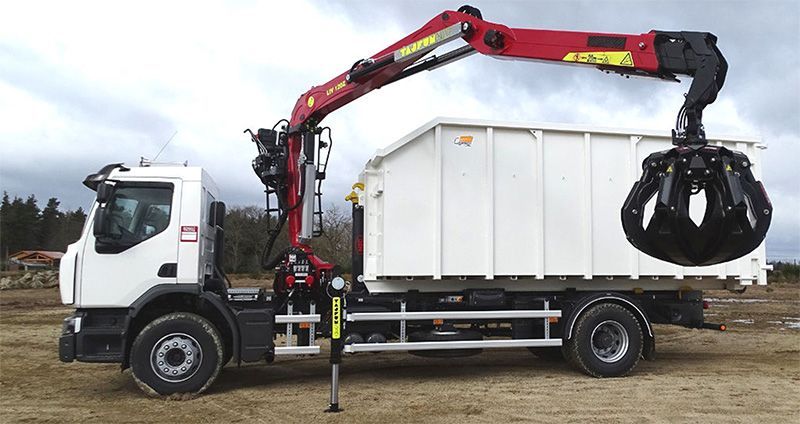 Truck with red crane arm lifting a black grapple claw over a white container in an outdoor setting.
