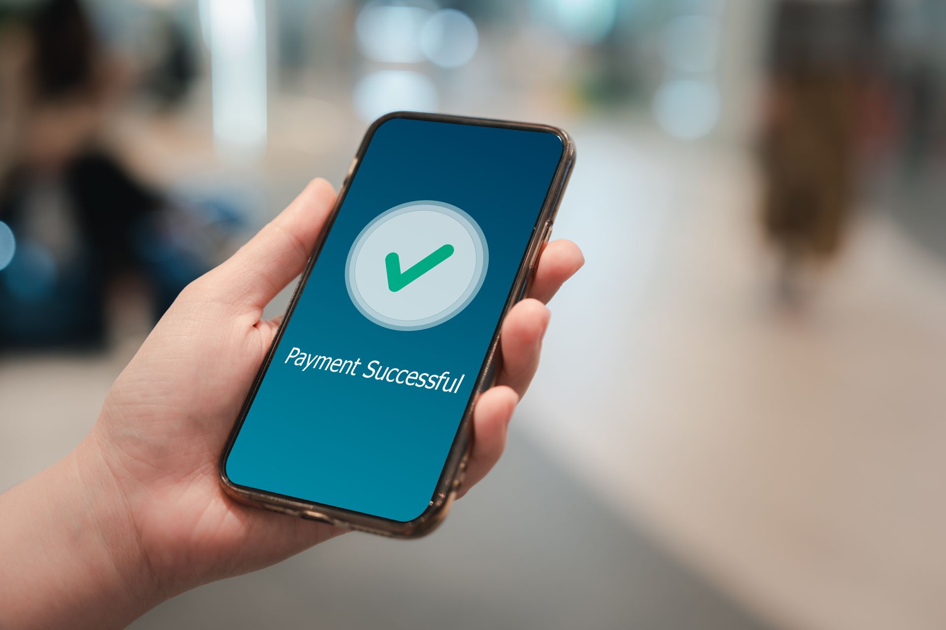 Hand holding a smartphone showing a green checkmark on a blue screen, with blurred background