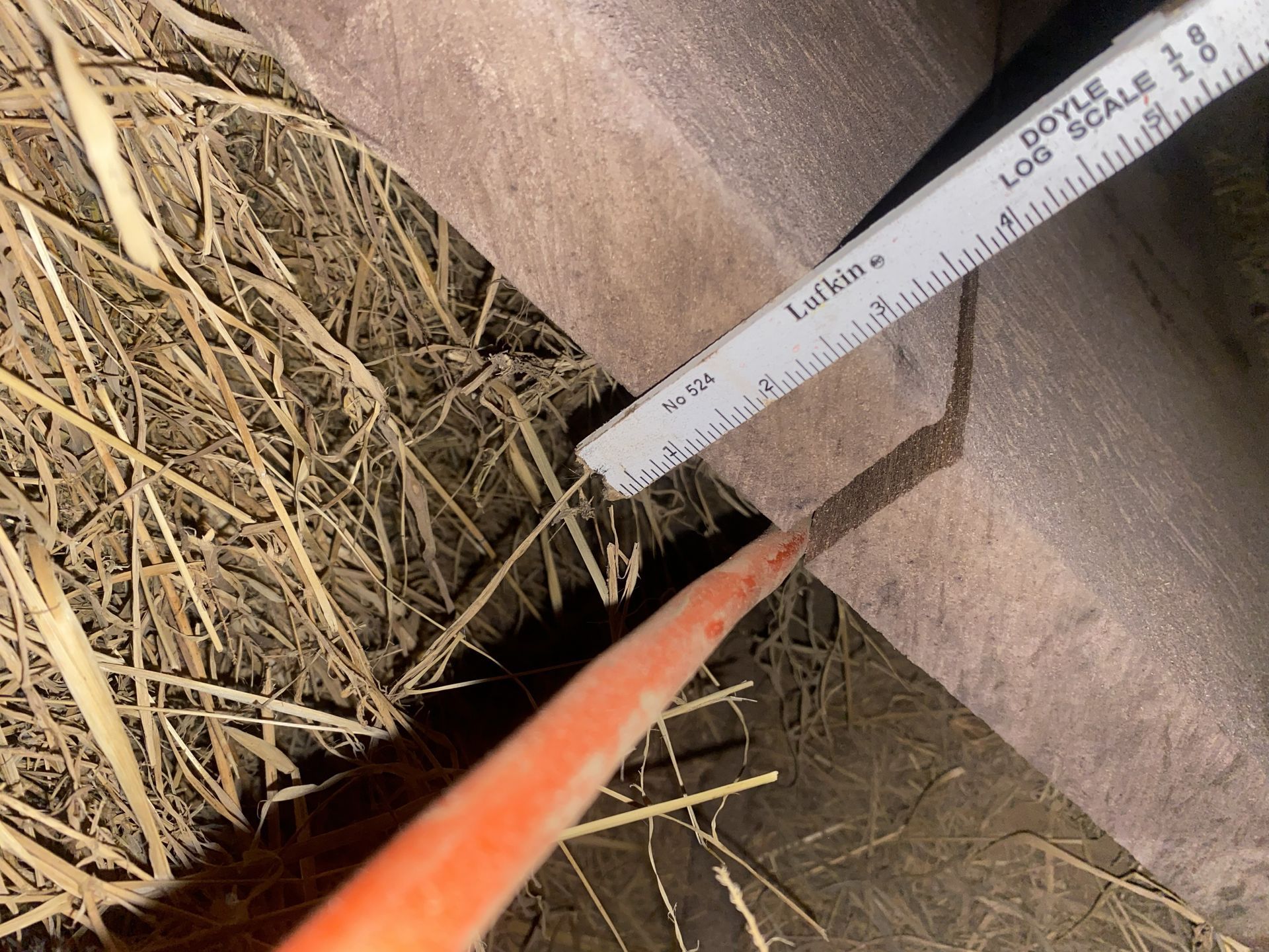A ruler and a bright orange object, likely a tool, measure a gap in a brown structure over straw.