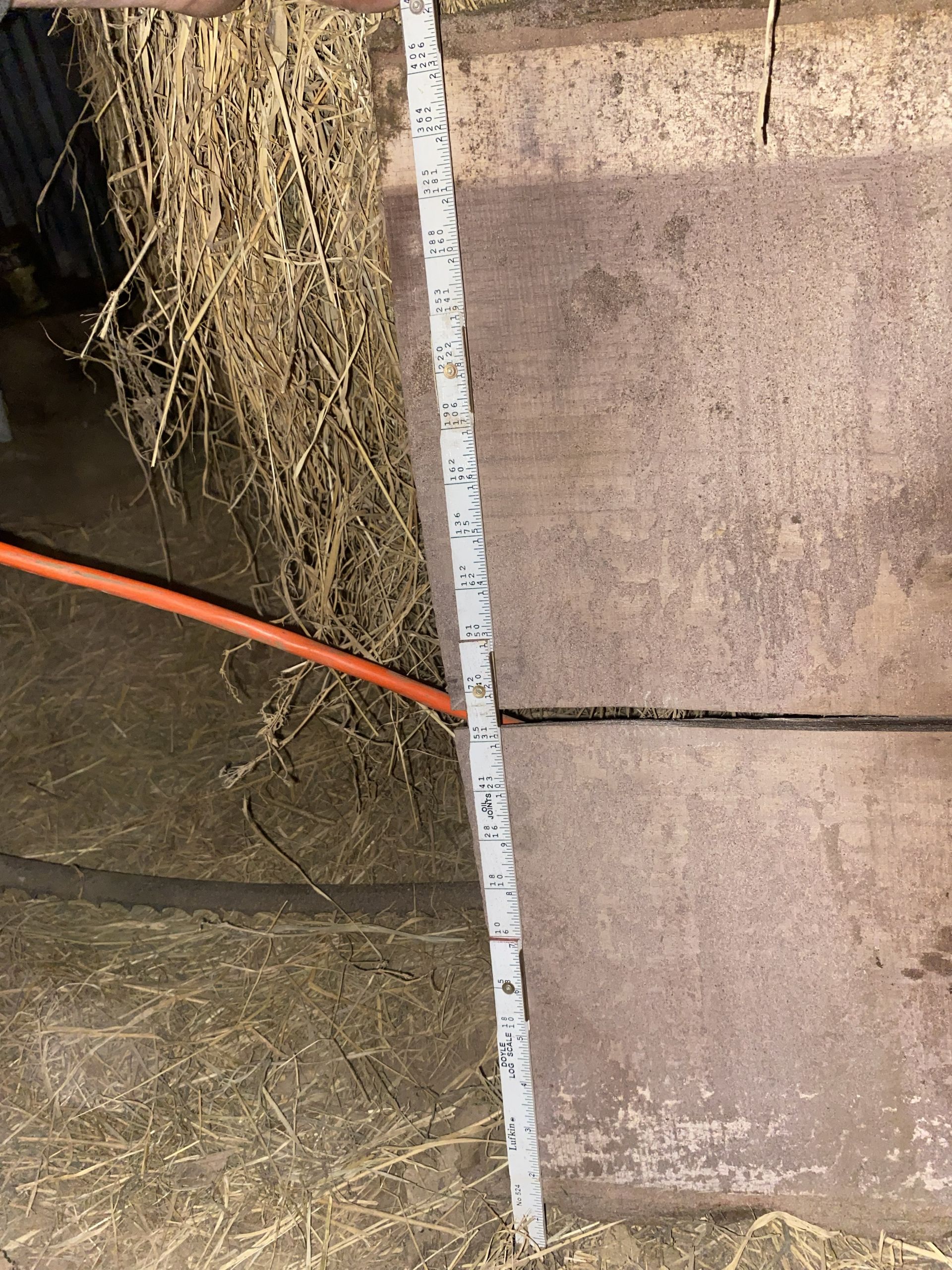 A measuring stick leaning against a large metal object in a dimly lit space with straw.