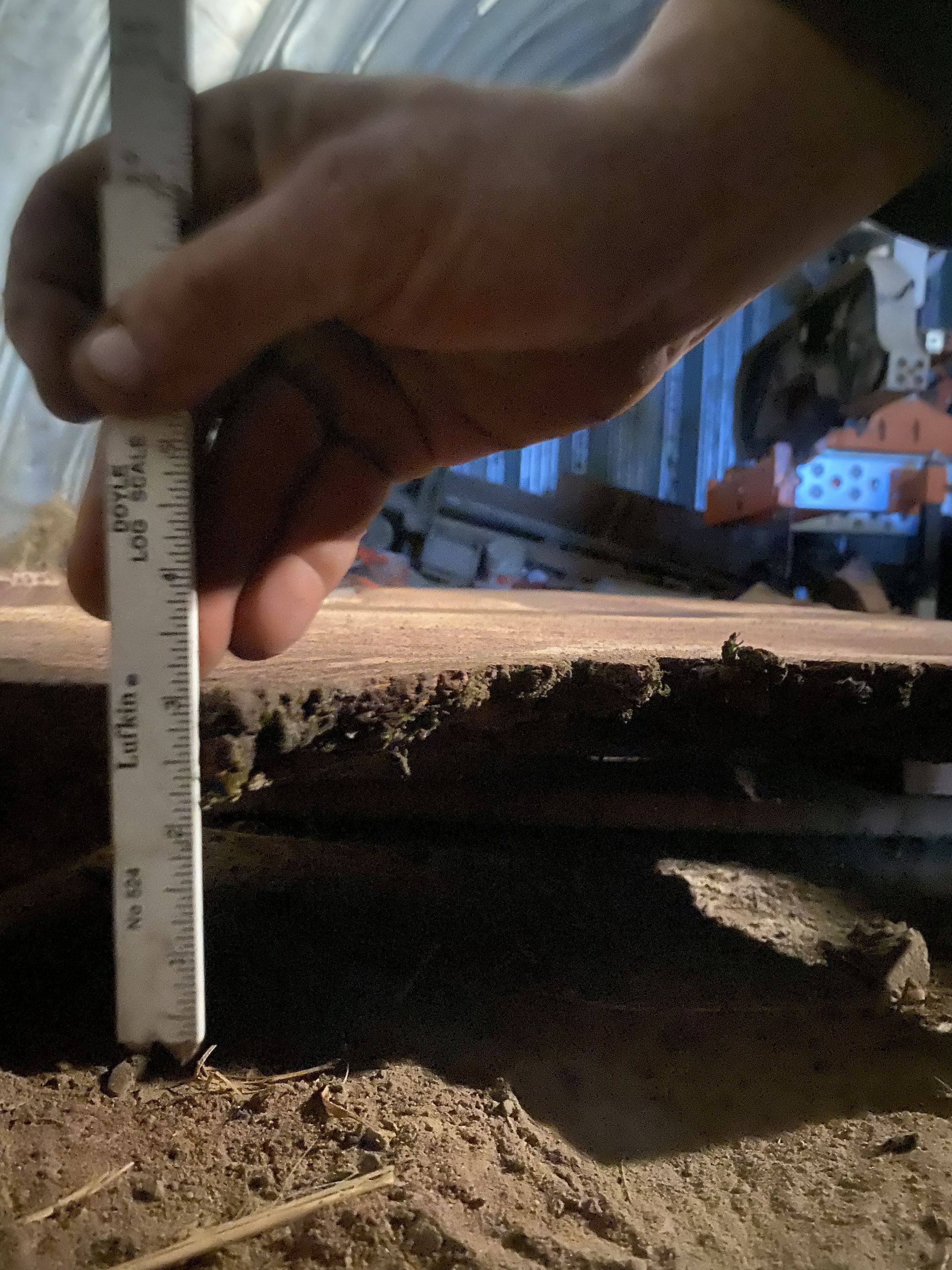 Hand using a ruler to measure the depth of dark, granular material. Wooden surface and machinery in the background.