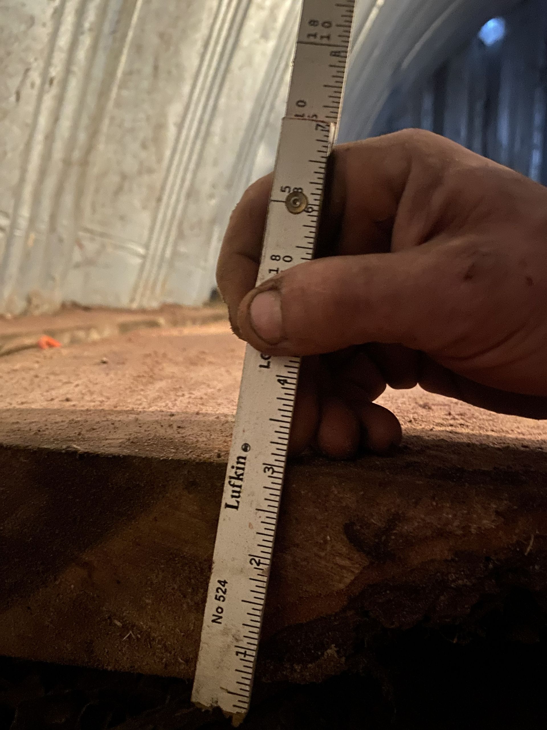 Hand holding a ruler measuring the thickness of a wooden slab in a dimly lit enclosure.